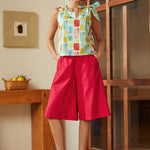 Woman wearing a colorful top and red shorts standing in a room with wooden furniture.
