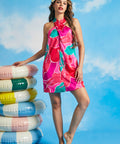 Woman wearing vibrant tropical floral halter dress beside stacked pastel pool floats