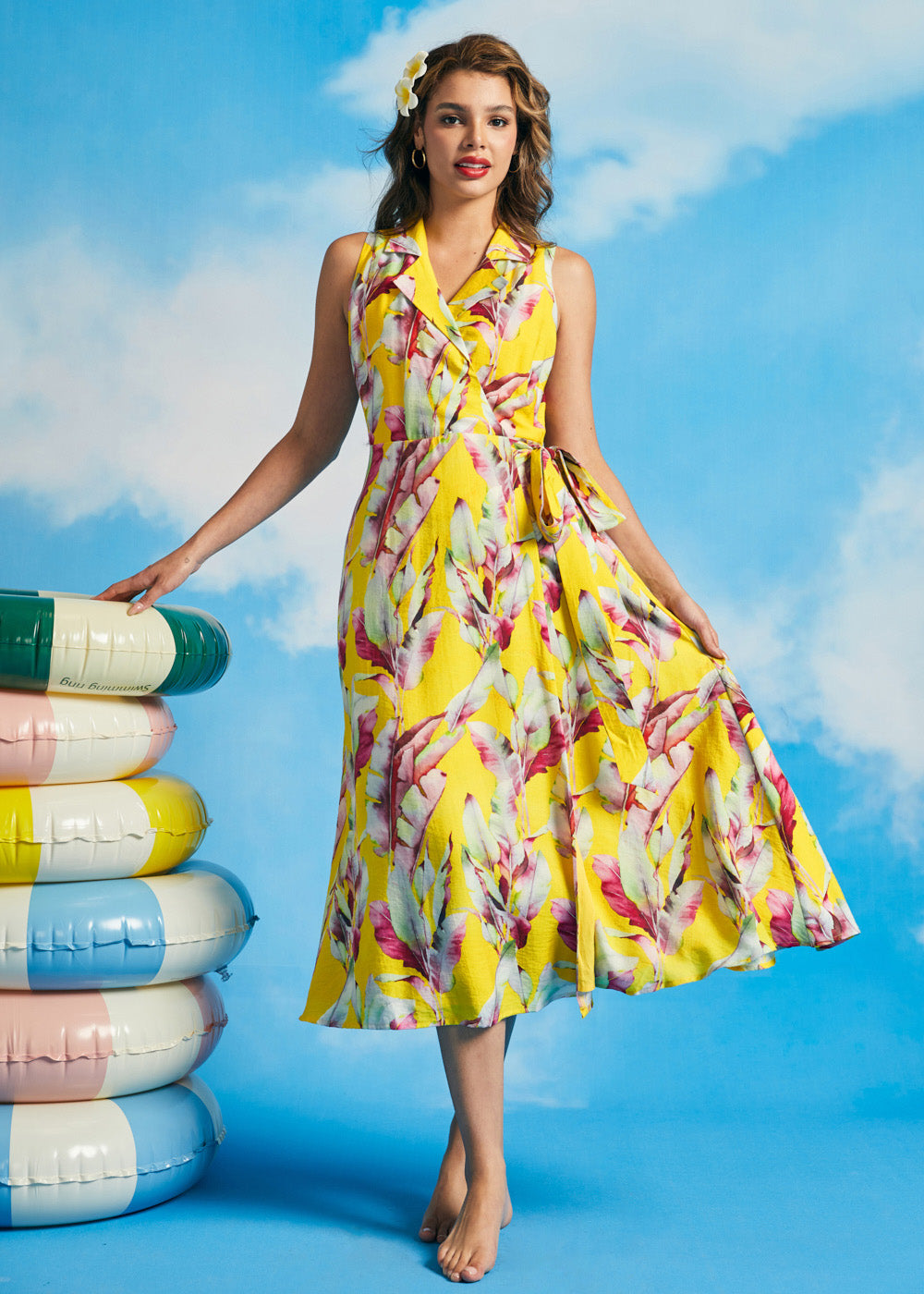 Woman wearing yellow tropical leaf print sleeveless wrap midi dress with tie waist and flowing skirt