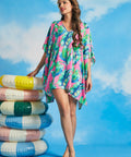 Model wearing pink tropical leaf print caftan cover-up with V-neckline and flowy silhouette beside inflatable pool rings