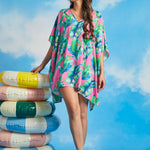 Model wearing pink tropical leaf print caftan cover-up with V-neckline and flowy silhouette beside inflatable pool rings