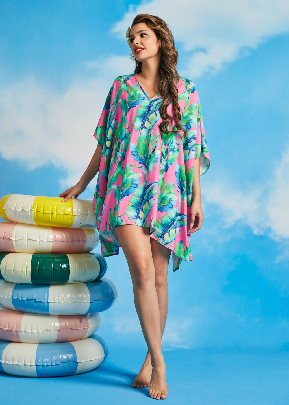 Model wearing pink tropical leaf print caftan cover-up with V-neckline and flowy silhouette beside inflatable pool rings