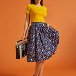 Woman in yellow top and novelty print skirt holding a vintage radio against an orange background