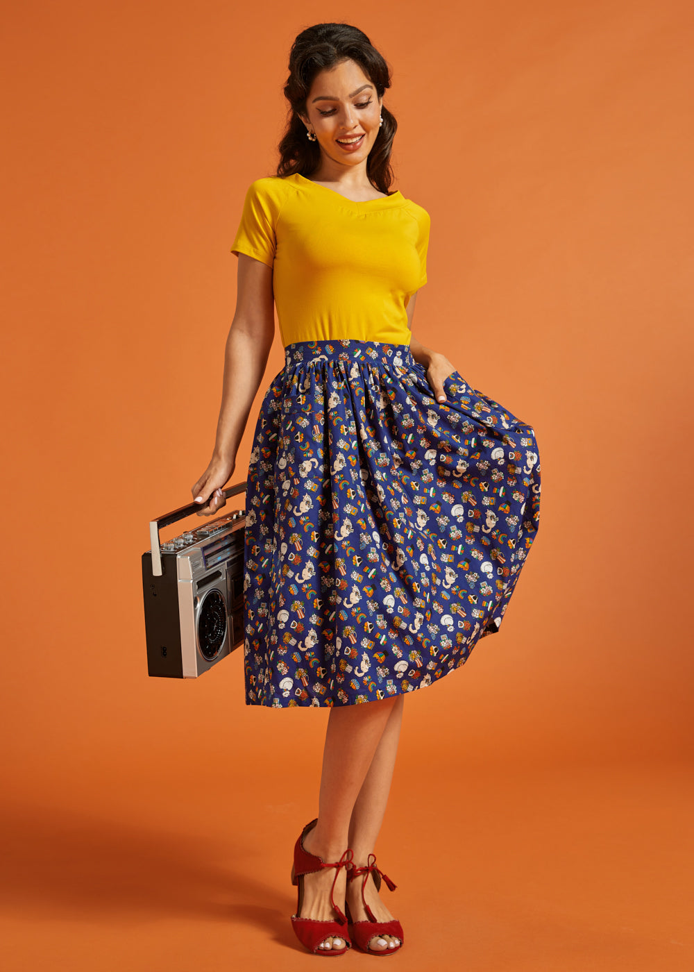 Woman in yellow top and novelty print skirt holding a vintage radio against an orange background