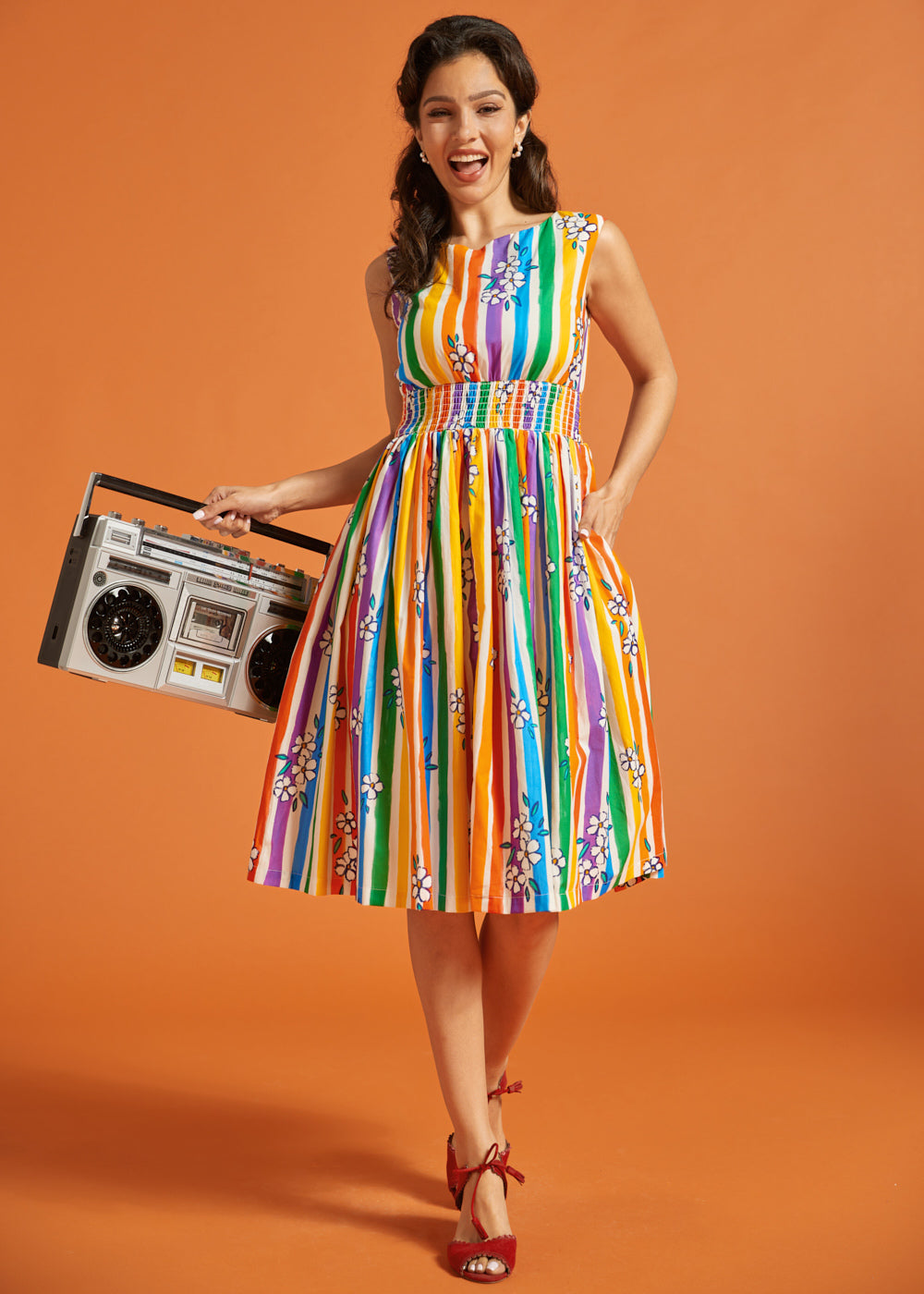 Woman in a sleeveless rainbow striped dress holding a retro boombox against an orange background