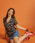 Woman wearing a rainbow patterned short sleeve shirt and denim skirt, sitting on the floor with red roller skates on