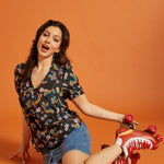Woman wearing a rainbow patterned short sleeve shirt and denim skirt, sitting on the floor with red roller skates on