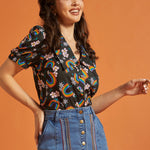 Woman wearing a vintage inspired colorful patterned top and blue jean skirt against an orange background
