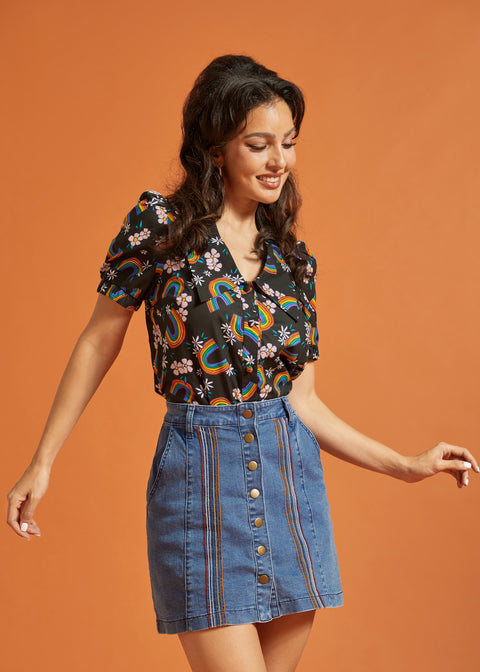 Woman wearing a rainbows and flower patterned top and denim skirt against an orange background