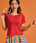 Woman wearing a red top and colorful skirt holding sunglasses against an orange background