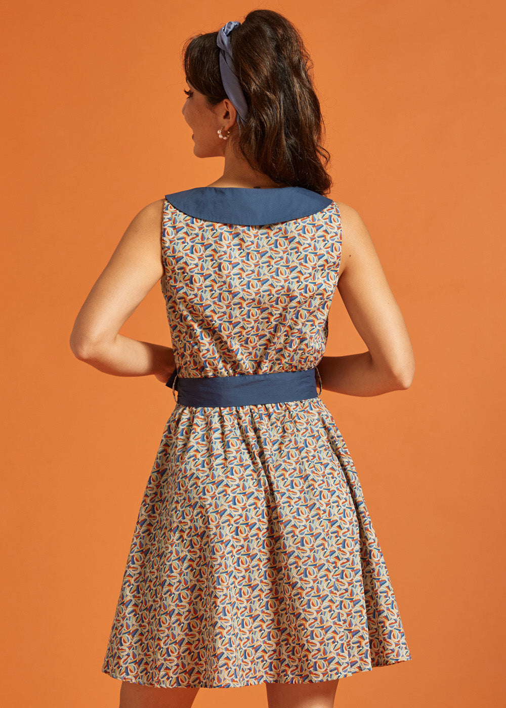 Back view of a retro-inspired sleeveless horseshoe print dress with a statement blue collar and coordinating tie belt.
