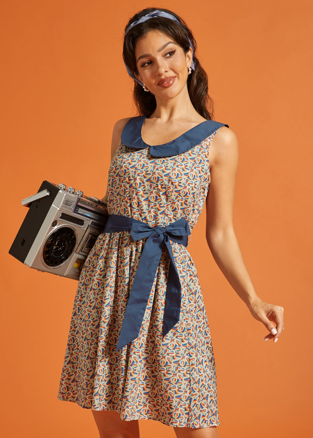 Woman carrying a boombox while wearing a retro fit-and-flare dress with a horseshoe print, oversized Peter Pan collar, and waist tie.