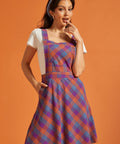 Woman wearing purple and orange plaid pinafore dress with square neckline and front pocket