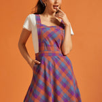 Woman wearing purple and orange plaid pinafore dress with square neckline and front pocket