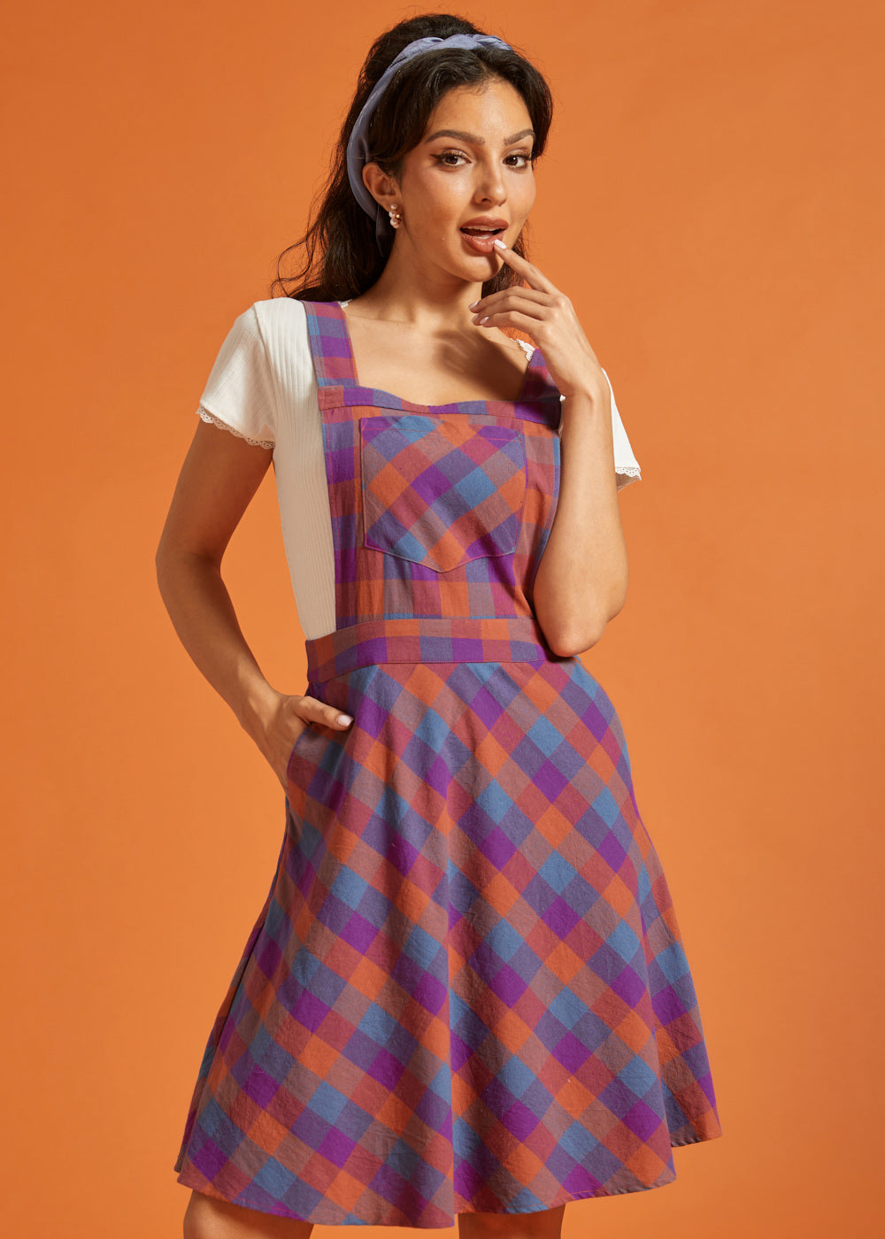 Woman wearing purple and orange plaid pinafore dress with square neckline and front pocket