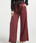 Woman wearing red tartan plaid wide leg pants with black sash belt and black top against a white studio background