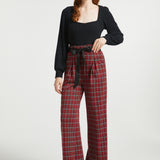 Full-length view woman wearing red tartan plaid wide leg pants with removable black bow belt and black heels against a white background