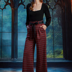 Woman wearing high-waisted red tartan plaid wide leg pants with black bow belt and black square-neck top in a gothic window setting