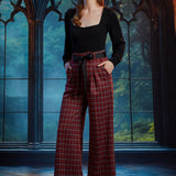 Woman wearing high-waisted red tartan plaid wide leg pants with black bow belt and black square-neck top in a gothic window setting