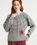 A woman wearing a gray knit sweater with a black spiderweb pattern and a red spider graphic on the front, paired with red plaid pants, against a light gray studio background.