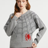 A woman wearing a gray knit sweater with a black spiderweb pattern and a red spider graphic on the front, paired with red plaid pants, against a light gray studio background.