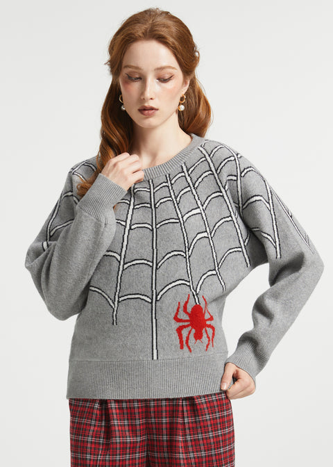 A woman wearing a gray knit sweater with a black spiderweb pattern and a red spider graphic on the front, paired with red plaid pants, against a light gray studio background.