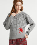 A woman with red hair wearing a gray knit sweater with a white spiderweb pattern and a red spider graphic on the front, paired with red and black plaid pants, against a light gray studio background.
