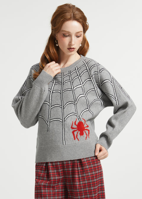 A woman with red hair wearing a gray knit sweater with a white spiderweb pattern and a red spider graphic on the front, paired with red and black plaid pants, against a light gray studio background.
