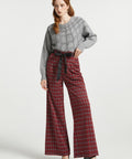 A woman modeling a gray knit sweater with spiderweb pattern and red plaid wide-leg trousers with a black tie belt, paired with black heels, against a light gray studio background.