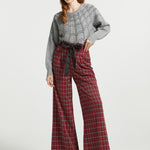 A woman modeling a gray knit sweater with spiderweb pattern and red plaid wide-leg trousers with a black tie belt, paired with black heels, against a light gray studio background.