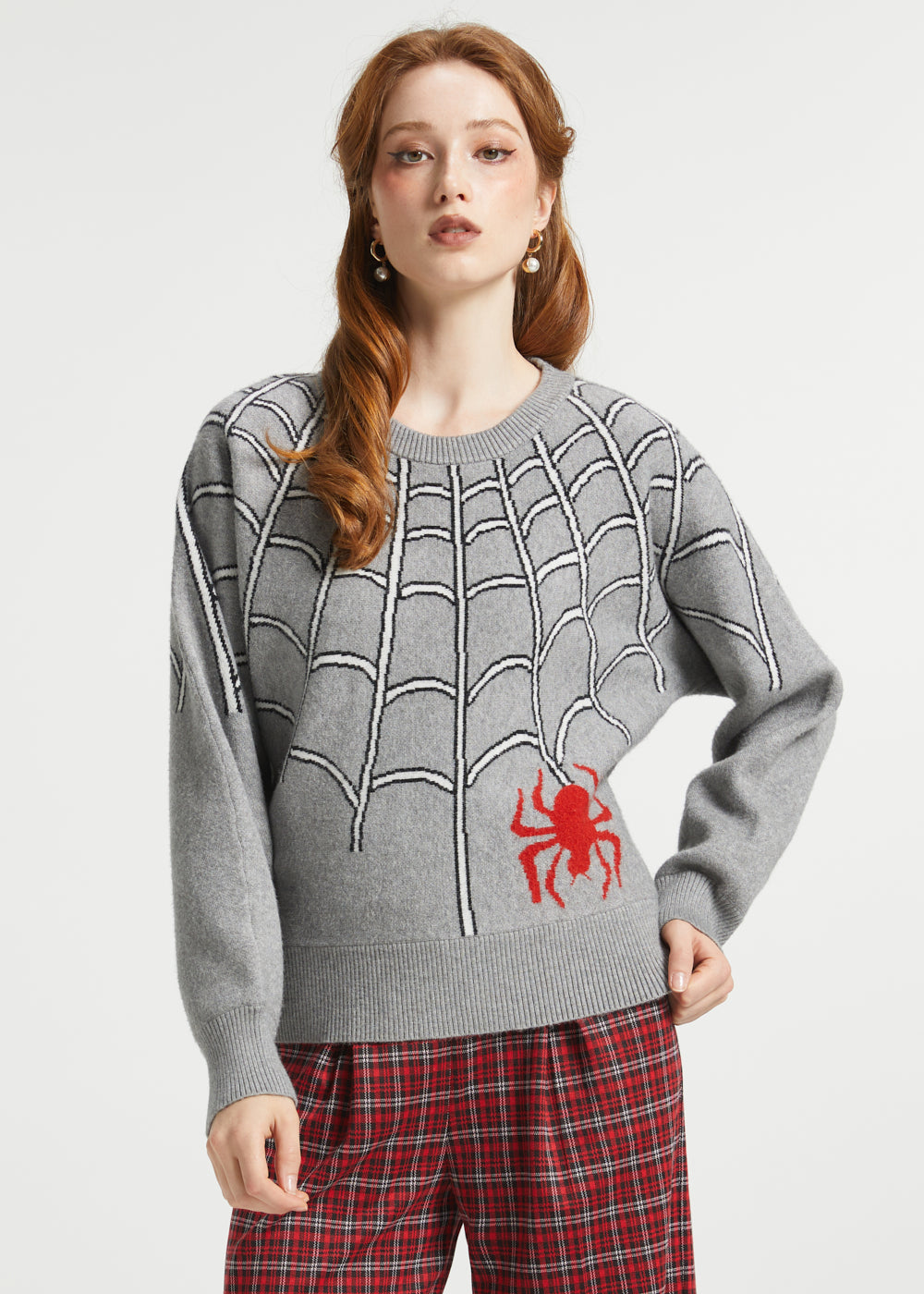 A woman modeling a gray knit sweater with a black spiderweb pattern and a red spider graphic on the lower right front, paired with red plaid pants against a light gray studio background.