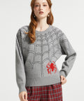 A woman modeling a gray knit sweater with a black spiderweb pattern and a red spider graphic on the lower right front, paired with red plaid pants against a light gray studio background.