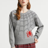 A woman modeling a gray knit sweater with a black spiderweb pattern and a red spider graphic on the lower right front, paired with red plaid pants against a light gray studio background.