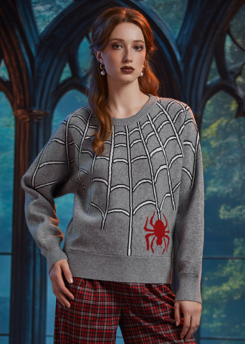 A woman wearing a gray knit sweater with a white spiderweb pattern and a red spider graphic on the lower front, paired with red plaid pants, posed in front of a dark gothic-style arched window.
