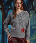 A woman wearing a gray knit sweater with a white spiderweb pattern and a red spider graphic on the lower front, paired with red plaid pants, posed in front of a dark gothic-style arched window.