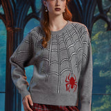 A woman wearing a gray knit sweater with a white spiderweb pattern and a red spider graphic on the lower front, paired with red plaid pants, posed in front of a dark gothic-style arched window.