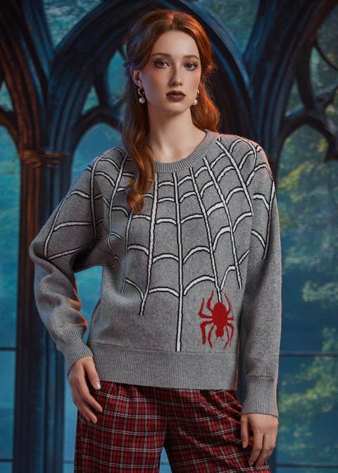 A woman wearing a gray knit sweater with a white spiderweb pattern and a red spider graphic on the lower front, paired with red plaid pants, posed in front of a dark gothic-style arched window.
