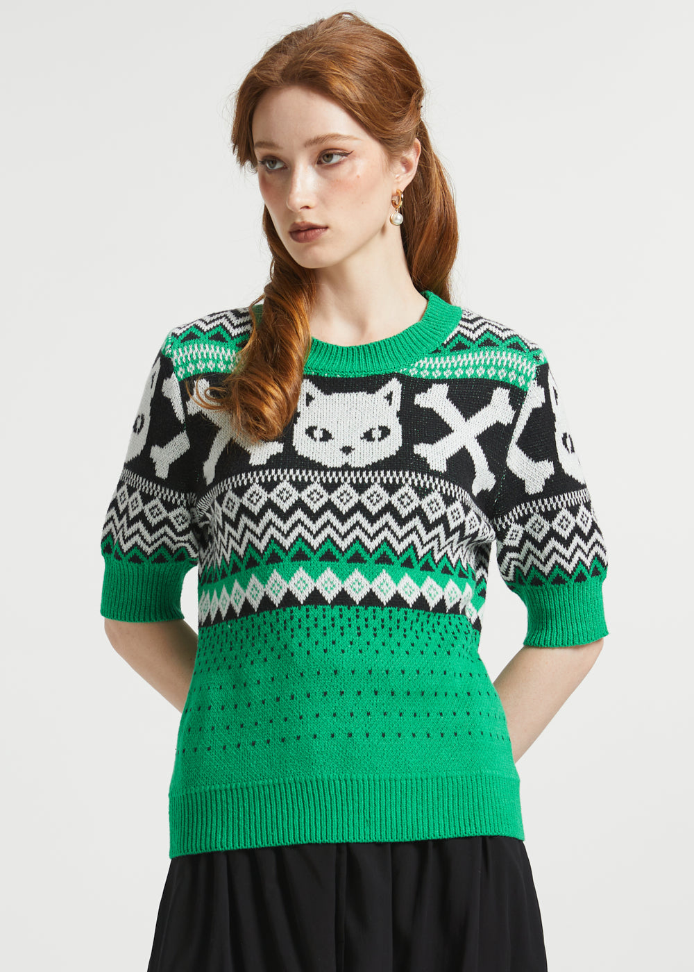 A woman wearing a green, black, and white patterned knit sweater with a cat face and skull-and-crossbones motif.