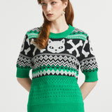 A woman wearing a green, black, and white patterned knit sweater with a cat face and skull-and-crossbones motif.