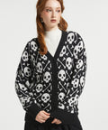 A woman wearing a black knit cardigan with a white skull and crossbones pattern, layered over a white ribbed turtleneck sweater and a black skirt, photographed against a light gray studio background.