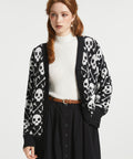 A woman modeling a black knit cardigan with a white skull and crossbones pattern, worn over a white ribbed turtleneck and a black skirt.