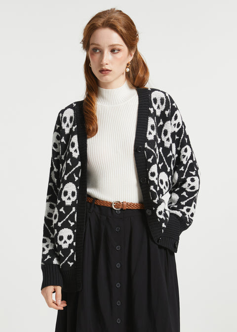 A woman modeling a black knit cardigan with a white skull and crossbones pattern, worn over a white ribbed turtleneck and a black skirt.