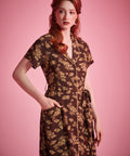 A woman modeling a dark brown short-sleeve dress with a beige floral pattern, standing against a solid pink studio background.