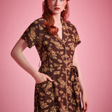 A woman modeling a dark brown short-sleeve dress with a beige floral pattern, standing against a solid pink studio background.