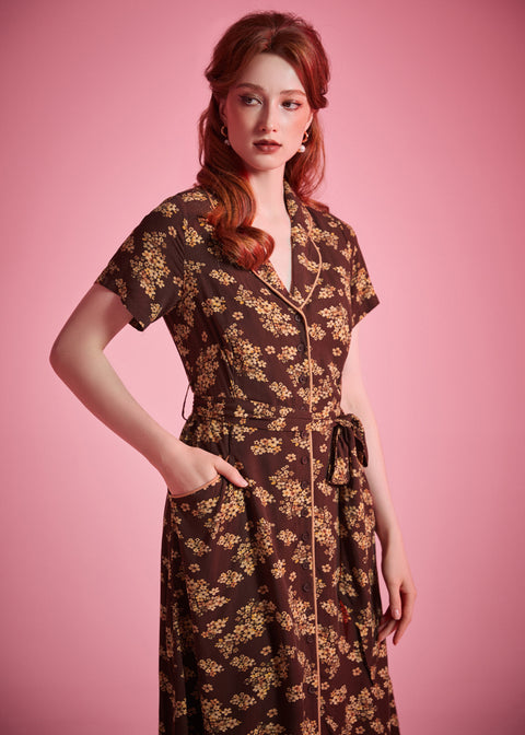 A woman modeling a dark brown short-sleeve dress with a beige floral pattern, standing against a solid pink studio background.