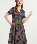 A woman modeling a short-sleeve plaid dress with a collared neckline, button front, and matching tie belt, shown against a plain light gray studio background.