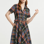 A woman modeling a short-sleeve plaid dress with a collared neckline, button front, and matching tie belt, shown against a plain light gray studio background.