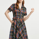 A woman modeling a short-sleeve plaid dress with a collared neckline, button front, and matching tie belt, shown against a plain light gray studio background.