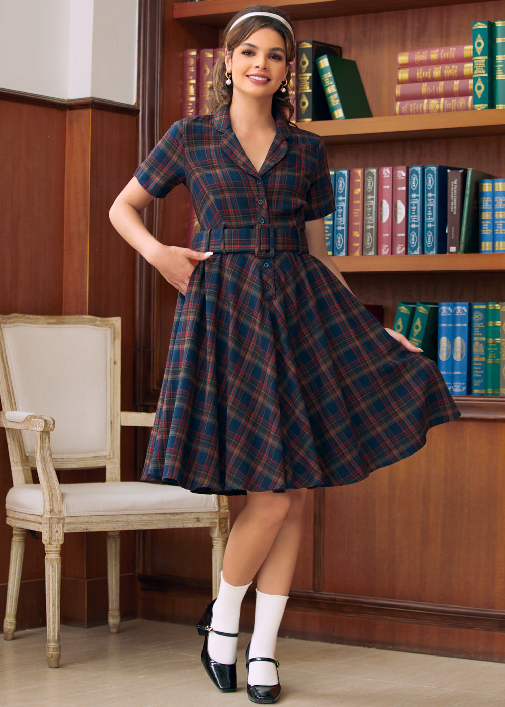 A woman models a short-sleeve plaid dress in a library setting with wooden bookshelves in the background. The dress features a navy blue, red, and green tartan pattern with a button-up front and a flared skirt.