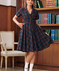 A woman models a short-sleeve plaid dress in a library setting with wooden bookshelves in the background. The dress features a navy blue, red, and green tartan pattern with a button-up front and a flared skirt.